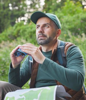 Vertical portrait of mature man holding binoculars and map while hiking alone in forest